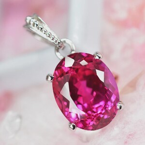 May include: A pendant featuring a large, oval-cut, vibrant fuchsia-pink gemstone set in a silver-toned metal prong setting. The top of the pendant includes a decorative, textured bail with a beaded edge, designed for attachment to a chain.