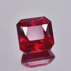 May include: A faceted, rectangular-cut ruby gemstone. The gem is a deep, saturated red colour with a reflective surface. The stone is displayed on a reflective surface, creating a mirror image of the ruby.