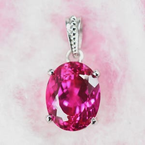 May include: An oval-cut, vibrant fuchsia-pink gemstone pendant set in a silver-toned metal prong setting. The pendant features a faceted surface that reflects light, attached to a small, textured silver-toned bail at the top for hanging.