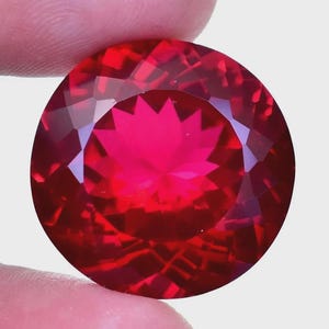 May include: A large, round, faceted gemstone with a deep red hue. The gem is held between two fingers, showcasing its brilliant cut and internal starburst pattern. The gem's facets reflect light, creating a dazzling display.