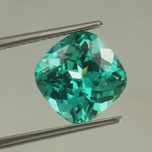 May include: A cushion-cut gemstone with a vibrant teal-green color and a faceted surface that reflects light. The gemstone is held by a pair of silver-toned metal tweezers, showcasing its transparent clarity and geometric shape against a neutral background.