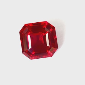 May include: A faceted, rectangular-cut ruby gemstone. The gem is a deep, vibrant red colour with sharp, clean edges and facets that reflect light. The gemstone is set against a white background.