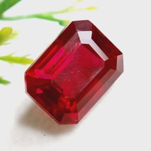 May include: A faceted, rectangular-cut ruby gemstone. The gem is a deep, vibrant red color with sharp, defined edges. The gemstone is displayed against a white background with a hint of green foliage.