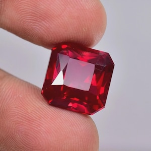 May include: A faceted, square-cut ruby gemstone. The gem is a deep, vibrant red color with multiple facets that reflect light. The ruby is held between two fingers against a plain background.