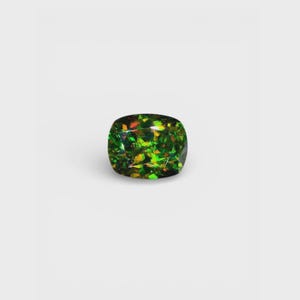 Puede incluir: Una piedra preciosa de corte cojín con un color base verde intenso y destellos iridiscentes vibrantes y multicolores en naranja, amarillo y verde lima en toda su superficie facetada, creando un juego de luz complejo.