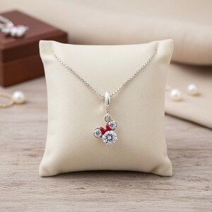 May include: A silver necklace with a pendant featuring a Minnie Mouse design. The pendant has a large, clear, round crystal as the face, with two smaller crystals for ears and a red bow. The necklace is displayed on a cream-colored pillow.