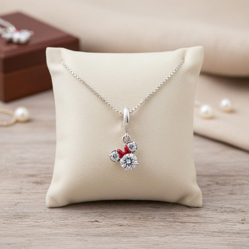 May include: A silver necklace with a pendant featuring a Minnie Mouse design. The pendant has a large, clear, round crystal as the face, with two smaller crystals for ears and a red bow. The necklace is displayed on a cream-colored pillow.
