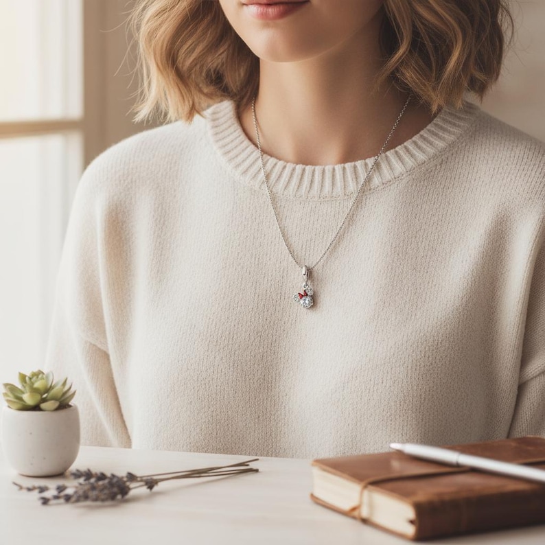 May include: A silver necklace with a pendant featuring a snowman design. The pendant has a red bow and is adorned with clear crystals. The person is wearing a cream-colored sweater.