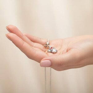 May include: A silver pendant necklace featuring a Minnie Mouse design. The pendant has a large, clear, round gemstone, two smaller clear gemstones, and a red gemstone. The necklace is held in a hand.