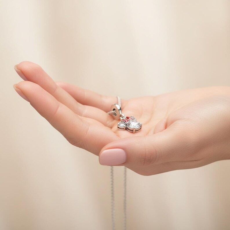 May include: A silver pendant necklace featuring a Minnie Mouse design. The pendant has a large, clear, round gemstone, two smaller clear gemstones, and a red gemstone. The necklace is held in a hand.