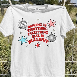 May include: A white short-sleeved t-shirt featuring a graphic print with the text DANCING IS EVERYTHING EVERYTHING EVERYTHING ELSE IS BULLSHIT in red letters, accompanied by two black and white disco balls and three stars in pink, blue, and red.