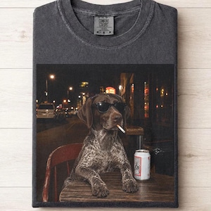 May include: A charcoal gray crew neck t-shirt featuring a graphic print of a brown and white spotted dog wearing black sunglasses, holding a cigarette in its mouth, and sitting at a wooden table next to a silver beverage can in a city setting.