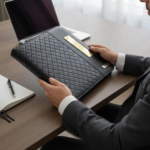 Op de afbeelding: Een zwarte leren portfolio met een ruitvormig patroon en een goudkleurige metalen plaat. De portfolio wordt vastgehouden door een persoon in een pak. Een laptop en notitieboek liggen op het houten bureau.