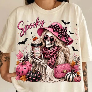 May include: A cream-colored t-shirt featuring a graphic of a skeleton wearing a pink hat with black polka dots, heart-shaped sunglasses, and a pink leopard print scarf. The skeleton holds a cup labeled Pumpkin Spice & Everything Nice.
