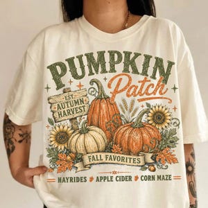 May include: An off-white t-shirt featuring a graphic print with the text PUMPKIN Patch, EST. AUTUMN HARVEST, FALL FAVORITES, HAYRIDES, APPLE CIDER, and CORN MAZE. The design includes orange pumpkins, yellow sunflowers, and autumn leaves.