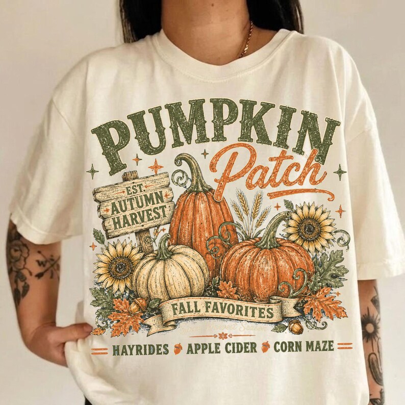 May include: An off-white t-shirt featuring a graphic print with the text PUMPKIN Patch, EST. AUTUMN HARVEST, FALL FAVORITES, HAYRIDES, APPLE CIDER, and CORN MAZE. The design includes orange pumpkins, yellow sunflowers, and autumn leaves.