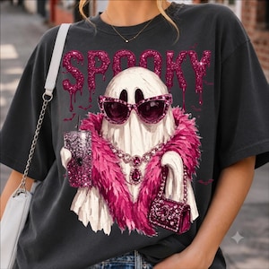 May include: A charcoal gray t-shirt featuring a graphic of a white ghost wearing pink cat-eye sunglasses, a pink feather boa, and a jeweled necklace. The ghost holds a glittery pink tumbler with a spiderweb design and a small pink handbag.