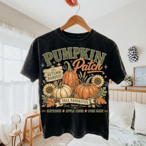 May include: A black short-sleeve t-shirt featuring a graphic print with the text PUMPKIN PATCH, EST. AUTUMN HARVEST, FALL FAVORITES, SINCE HARVEST SEASON, HAYRIDES, APPLE CIDER, and CORN MAZE, alongside illustrations of pumpkins, sunflowers, and leaves.