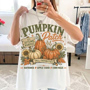 May include: A white t-shirt featuring a graphic print with the text PUMPKIN Patch, EST. AUTUMN HARVEST, FALL FAVORITES, HAYRIDES, APPLE CIDER, and CORN MAZE. The design includes illustrations of orange pumpkins, sunflowers, and autumn leaves.