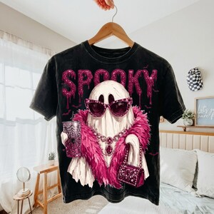 May include: A black mineral-washed t-shirt featuring a graphic of a white ghost wearing pink glittery sunglasses, a pink feather boa, and a jeweled necklace. The ghost holds a glittery pink tumbler and a matching handbag. The word SPOOKY is printed above.