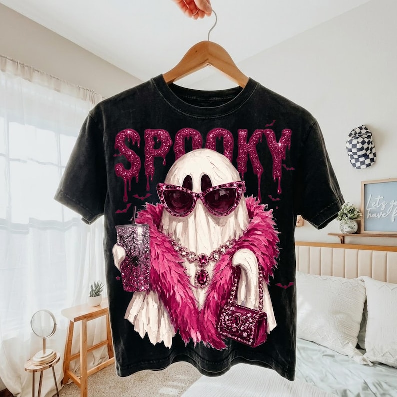 May include: A black mineral-washed t-shirt featuring a graphic of a white ghost wearing pink glittery sunglasses, a pink feather boa, and a jeweled necklace. The ghost holds a glittery pink tumbler and a matching handbag. The word SPOOKY is printed above.