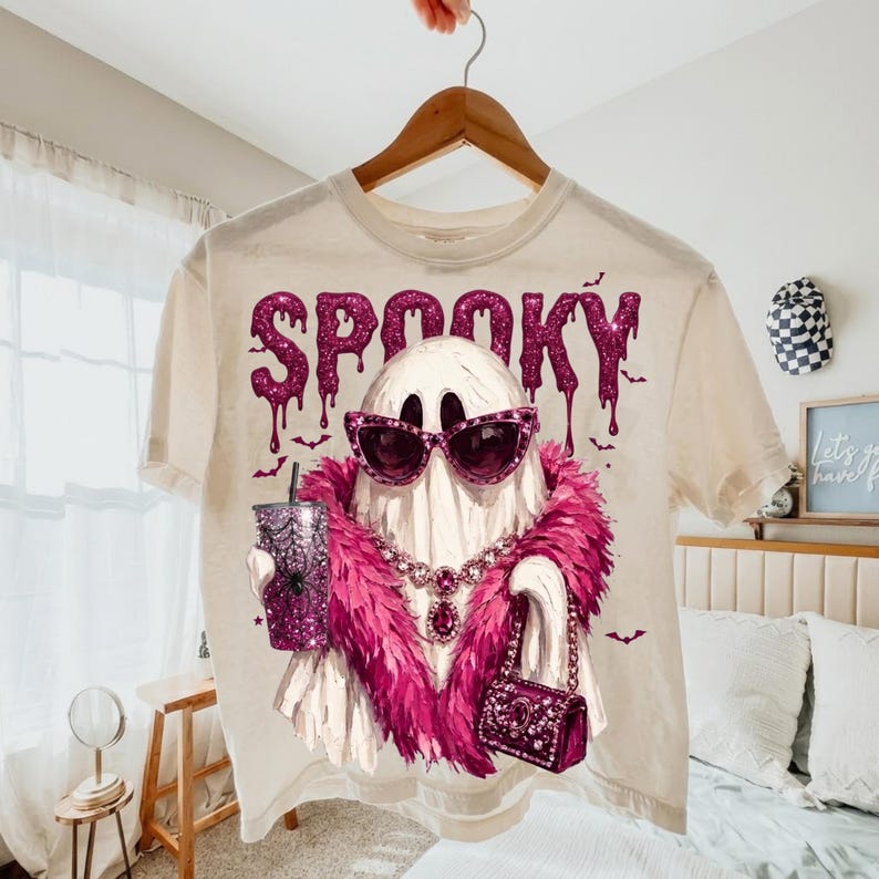 May include: A cream-colored t-shirt featuring a graphic of a white ghost wearing pink cat-eye sunglasses, a pink feather boa, and a jeweled necklace. The ghost holds a glittery pink tumbler and a matching handbag. The word SPOOKY is printed above.