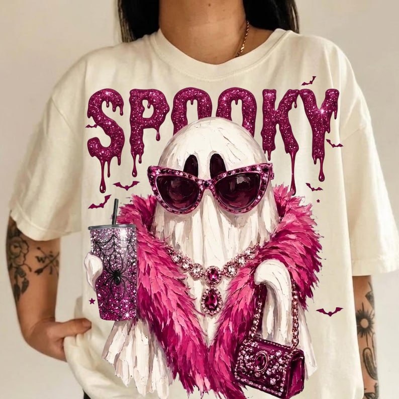 May include: An off-white t-shirt featuring a graphic of a white ghost wearing pink cat-eye sunglasses, a pink feather boa, and a jeweled necklace. The ghost holds a glittery pink tumbler with a spiderweb design and a small pink handbag.