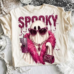 May include: A cream-colored t-shirt featuring a graphic of a ghost wearing pink cat-eye sunglasses, a pink feather boa, and a jeweled necklace. The ghost holds a glittery pink tumbler and a small handbag. The word SPOOKY is printed in pink glitter.
