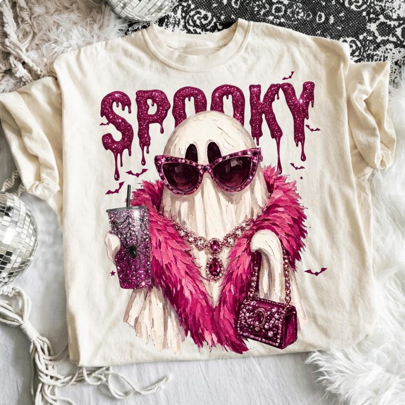 May include: A cream-colored t-shirt featuring a graphic of a ghost wearing pink cat-eye sunglasses, a pink feather boa, and a jeweled necklace. The ghost holds a glittery pink tumbler and a small handbag. The word SPOOKY is printed in pink glitter.
