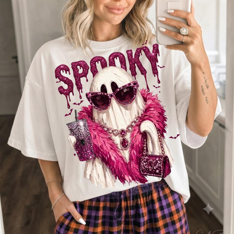 May include: A white t-shirt featuring a graphic of a ghost wearing pink glittery sunglasses, a pink feather boa, and a jeweled necklace. The ghost holds a glittery tumbler and a handbag. The word SPOOKY is printed in dripping pink glitter text.