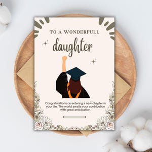 Puede incluir: Una tarjeta de felicitación de graduación con el texto "TO A WONDERFULL daughter". La tarjeta presenta una ilustración de un graduado con toga y birrete. La tarjeta está sobre un plato de madera con un sobre marrón.