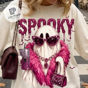 May include: A cream-colored sweatshirt featuring a graphic of a white ghost wearing pink cat-eye sunglasses, a pink feather boa, and a jeweled necklace. The ghost holds a glittery pink tumbler with a spiderweb design and a small pink handbag.