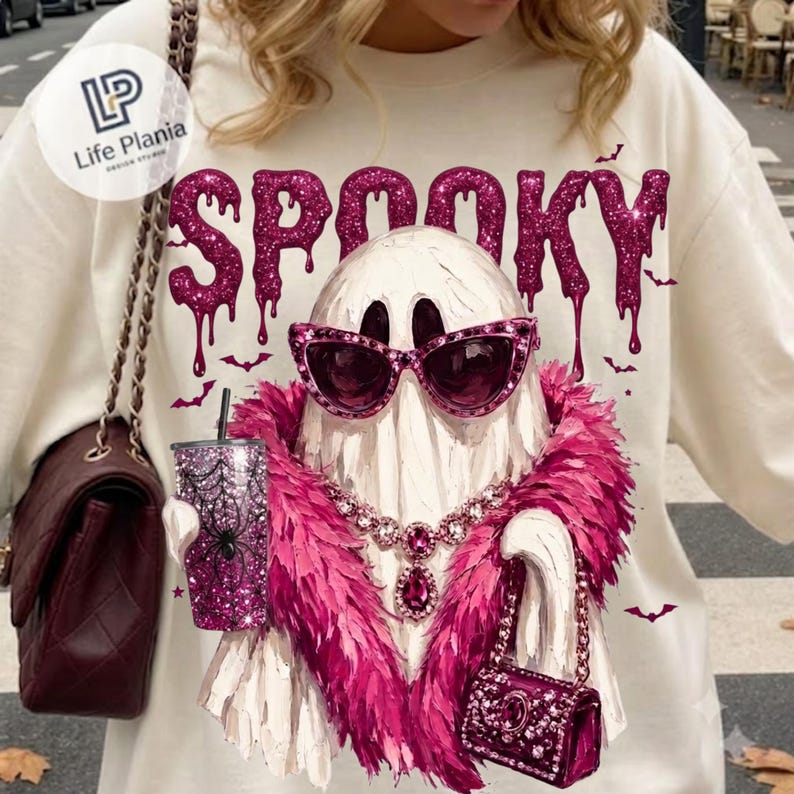 May include: A cream-colored sweatshirt featuring a graphic of a white ghost wearing pink cat-eye sunglasses, a pink feather boa, and a jeweled necklace. The ghost holds a glittery pink tumbler with a spiderweb design and a small pink handbag.
