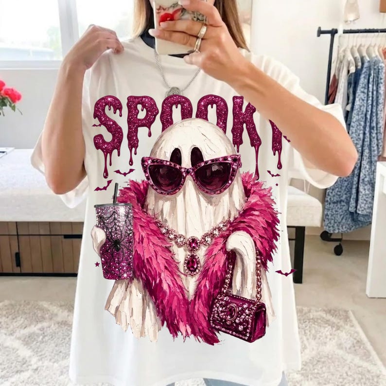 May include: A white t-shirt featuring a graphic of a ghost wearing pink cat-eye sunglasses, a pink feather boa, and a pink necklace. The ghost holds a pink glittery tumbler with a spiderweb design and a small pink handbag. The text SPOOKY is above.