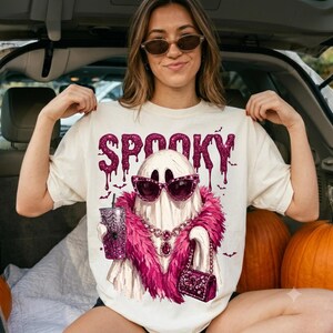 May include: A cream-colored t-shirt featuring a graphic of a ghost wearing pink cat-eye sunglasses, a pink feather boa, and a necklace. The ghost holds a glittery pink tumbler and a matching handbag. The word SPOOKY is printed in pink glitter above.