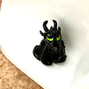Könnte beinhalten: Kleine, schwarze Drachenfigur mit leuchtend grünen Augen. Der Drache hat kleine Hörner und sitzt auf einer weißen Oberfläche. Die Figur ist detailliert und scheint handgefertigt zu sein.