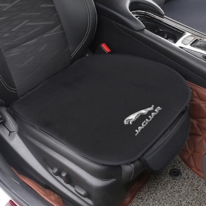 Puede incluir: Cojín negro para asiento de coche con el logotipo de Jaguar en plata. El cojín está colocado sobre un asiento de coche negro. El asiento tiene un ribete de cuero negro y una hebilla de cinturón de seguridad. El cojín está diseñado para el asiento.