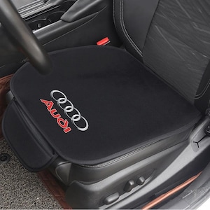 May include: Black car seat cover featuring the Audi logo in red and silver. The cover is placed on a black car seat. The Audi logo is on the lower right side of the seat cover.