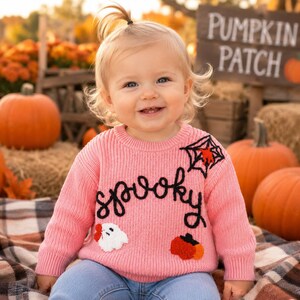 May include: A light pink long-sleeve knit sweater featuring the word "spooky" in black script, a black spiderweb with a red spider, a white ghost with a red bow, and an orange pumpkin. The sweater has a crew neckline and ribbed cuffs.