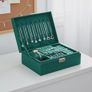 May include: A dark green jewelry box with an open lid, revealing various necklaces, rings, earrings, and a gold wristwatch. The box is lined with a soft, green fabric and has a gold-colored clasp.