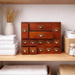 May include: A wooden storage cabinet with multiple drawers, featuring antique brass-toned handles. The cabinet is stained a warm brown and sits on a wooden shelf, alongside white towels, a vase with dried plants, and other storage items.
