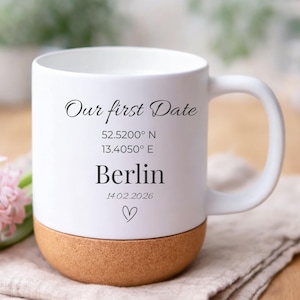 Könnte beinhalten: Weißer Keramikbecher mit Korkboden, mit dem Text "Our first Date", Koordinaten, der Stadt "Berlin" und dem Datum 14.02.2026, mit einem Herzsymbol. Der Becher steht auf einem beigen Tuch.