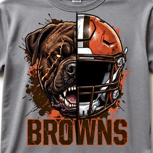 Könnte beinhalten: Graues T-Shirt mit geteiltem Design: auf der einen Seite ein knurrender brauner Hund und auf der anderen Seite ein orangefarbener Footballhelm. Das Wort "BROWNS" ist in großen orangefarbenen Buchstaben gedruckt. Das Design hat einen Used-Look.