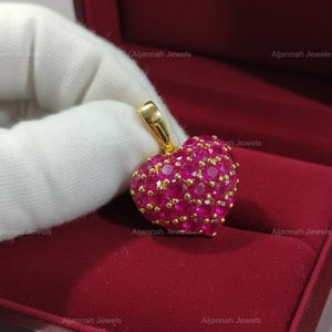 May include: A heart-shaped pendant featuring numerous small, round, vibrant pinkish-red gemstones set closely together in a gold-toned metal frame. The pendant includes a gold-toned bail at the top for attachment to a chain or necklace.