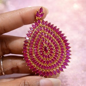 May include: A gold-toned pendant with a teardrop design, densely covered in small, vibrant pink gemstones. The pendant has a central, larger gemstone and is held by a hand against a soft pink background. The jewelry piece is from Aljannah Jewels.