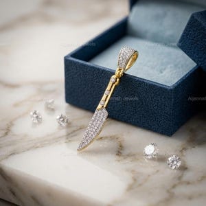 May include: A gold pendant shaped like a knife, encrusted with small, sparkling stones. The pendant rests on a marble surface next to a blue jewelry box. Several loose gemstones are scattered nearby. The brand name "Aljannah Jewels" is visible.