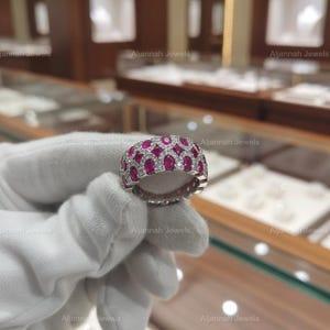 May include: A silver ring with a repeating pattern of oval-shaped red gemstones and small white gemstones. The ring is held by a gloved hand in front of a jewelry display case. The brand name "Aljannah Jewels" is visible in the background.