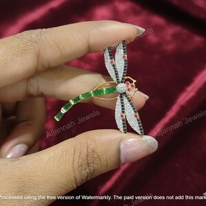 Può includere: Spilla a forma di libellula con metallo dorato, dettagli verde smeraldo, nero, bianco e rosso. Le ali sono decorate con pietre nere e bianche, e il corpo presenta pietre rettangolari verdi. La spilla è tenuta su uno sfondo bordeaux.