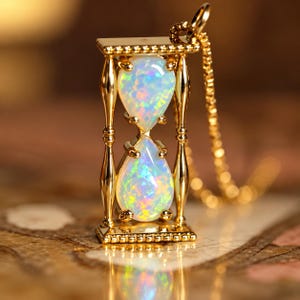 May include: A gold-toned pendant shaped like an hourglass, featuring two pear-shaped iridescent opals stacked vertically within a metallic frame. The pendant is attached to a delicate gold-toned chain, showcasing a polished finish.