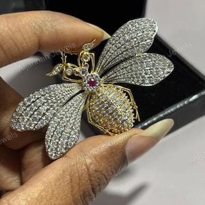 May include: A gold and silver bee brooch. The wings are covered in small, clear stones, and the body is gold with a central red stone. The brooch is held against a dark background.